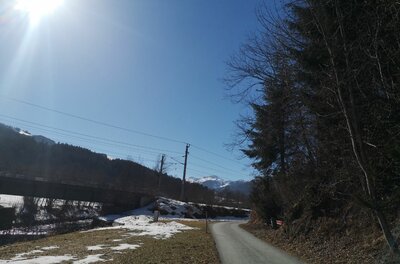 Asphaltierter Weg neben einer Bahnbrücke und verschneiten Wiesen unter blauem Himmel.