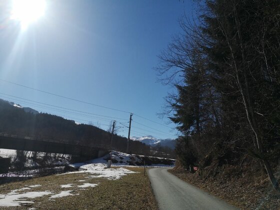 Asphaltierter Weg neben einer Bahnbrücke und verschneiten Wiesen unter blauem Himmel.