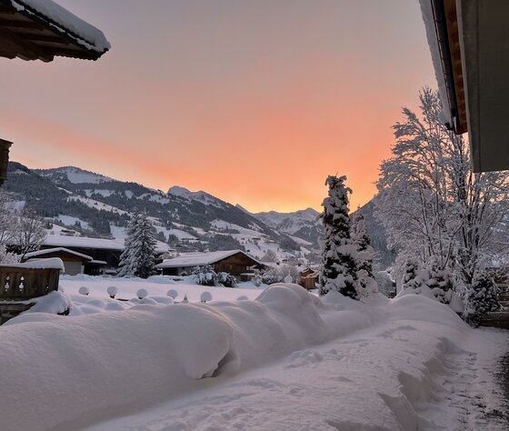 Ausblick Aurach im Winter Verschneite Alpendorf-Landschaft mit Holzhäusern und Bergen vor einem orangefarbenen Abendhimmel.