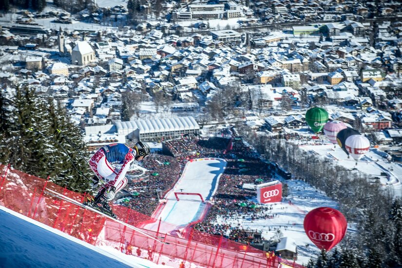 Skirennfahrer in Abfahrtshocke auf einer steilen Piste oberhalb eines Zielraums mit Zuschauermengen.