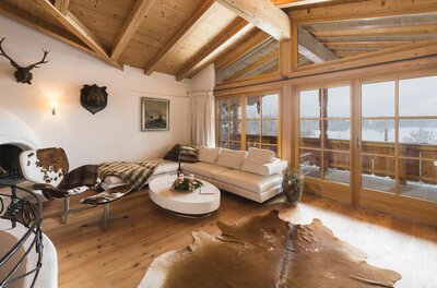 Rustikaler Wohnraum mit weißem Sofa, Holzboden, Fellteppich und großen Glasfronten mit Blick auf eine Schneelandschaft.