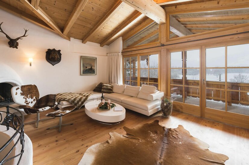 Rustikaler Wohnraum mit weißem Sofa, Holzboden, Fellteppich und großen Glasfronten mit Blick auf eine Schneelandschaft.