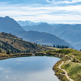 Ein klarer Bergsee spiegelt die umliegenden Berge und Wälder wider, während die Sonne den Herbsthimmel erhellt.