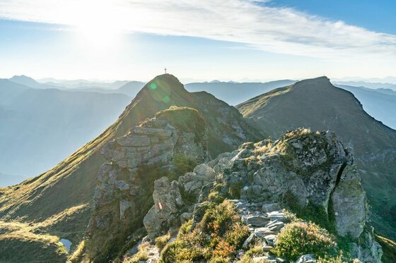 Felsiger Gebirgsgrat mit Gipfelkreuz im Gegenlicht vor weiteren Bergketten.