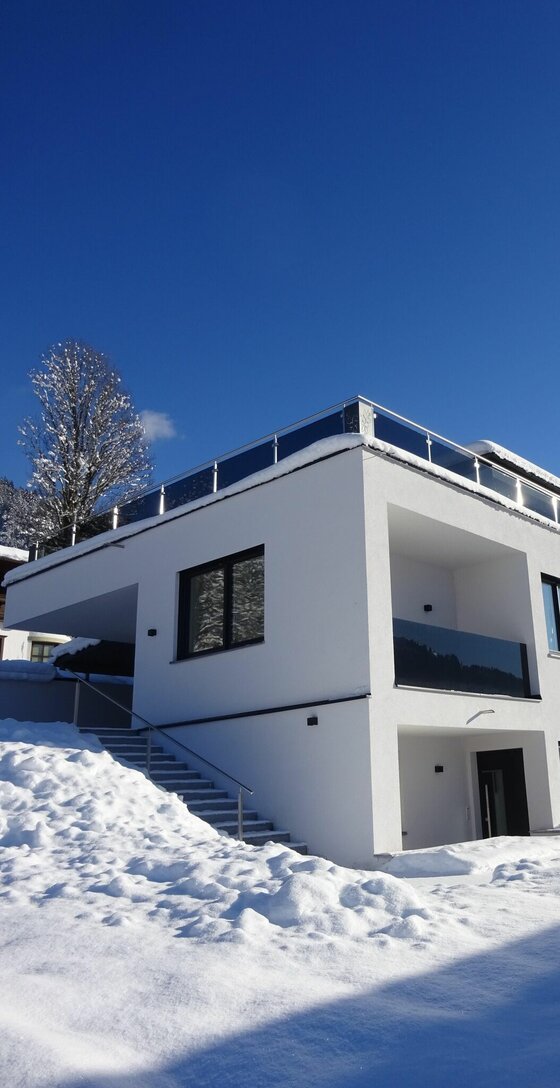 Modernes weißes Apartmentgebäude mit Flachdach und Glasbalkonen im Schnee unter blauem Himmel.