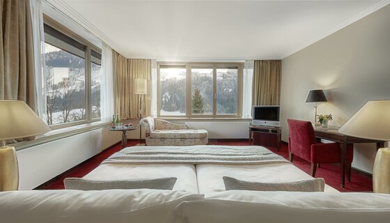 Hotelzimmer mit Doppelbett, Schreibtisch und Blick durch große Fenster auf eine verschneite Berglandschaft.