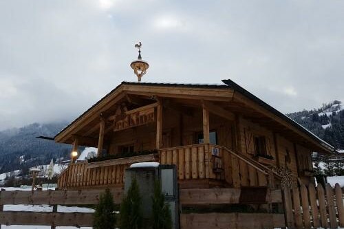 Holzhütte mit Terrasse und Wetterhahn vor verschneiten Bergen unter bewölktem Himmel.