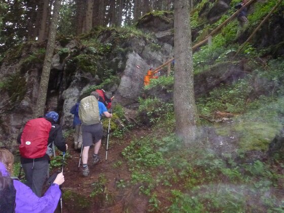 Wandergruppe mit Rucksäcken und Stöcken auf einem steilen Waldpfad an Felsen vorbei.
