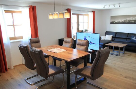 Moderner Wohn- und Essbereich mit Holztisch, Polsterstühlen, dunklem Sofa und Fernseher.
