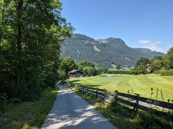 Asphaltierter Wanderweg neben einer grünen Wiese mit Holzzaun und bewaldeten Bergen im Hintergrund.