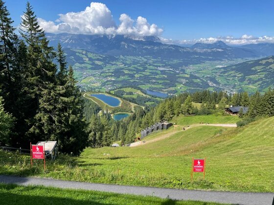 Blick von einem Grashang auf Bergseen und ein bewaldetes Tal vor einem Gebirgsmassiv unter Wolken.
