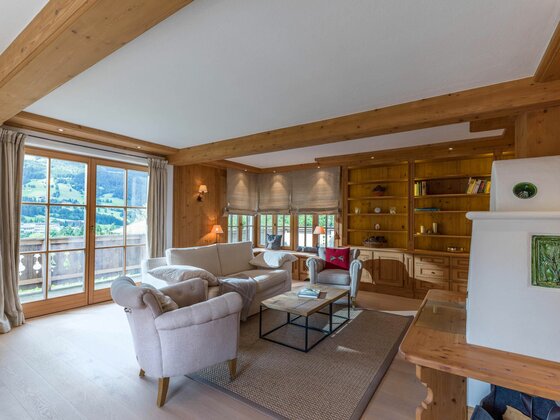 Wohnzimmer mit hellen Polstermöbeln, Holzwänden, Einbauregalen und Blick durch eine Glastür auf die Berge.