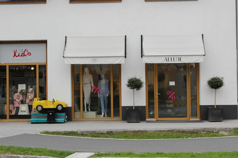 Außenansicht des Bekleidungsgeschäfts Allude mit Schaufenstern, Markisen und einem gelben Kinder-Spielauto.