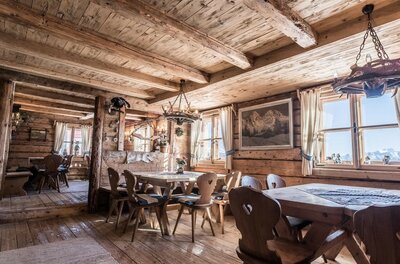 Rustikale Gaststube aus hellem Holz mit Holztischen, Stühlen mit Herz-Ausschnitt und Geweih-Leuchtern.