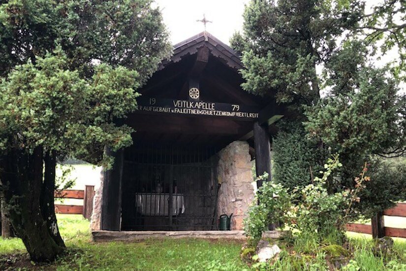 Eine kleine Kapelle, umgeben von grünen Bäumen und Pflanzen. Der Eingang ist mit einem Gitter versehen und ein Kreuz befindet sich auf dem Dach.