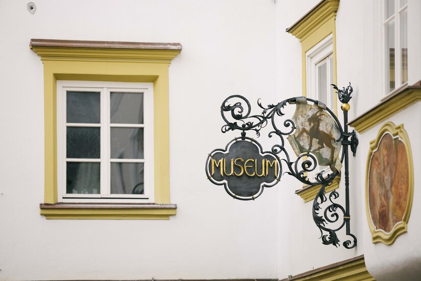 Ein schmiedeeisernes Schild mit der Aufschrift Museum und einem Steinbock-Wappen an einer weißen Hauswand.