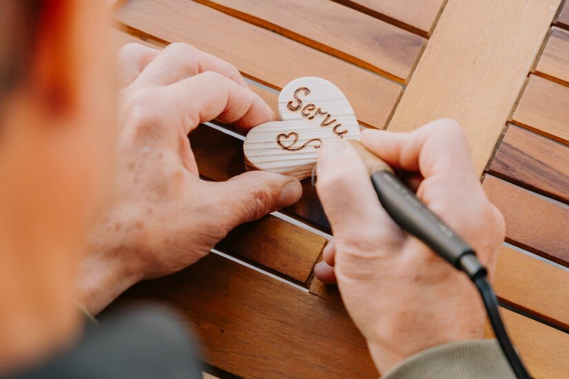 Hände brennen mit einem Brandmalkolben den Schriftzug Servus in ein kleines Holzherz.