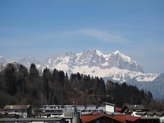 Blick über Hausdächer und einen Nadelwald auf ein schneebedecktes Felsmassiv unter blauem Himmel.