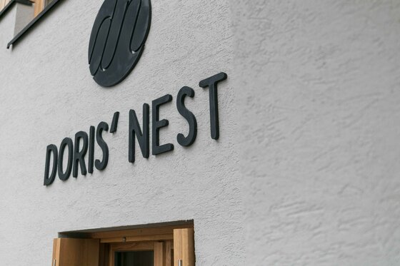Schwarzer Schriftzug Doris' Nest an einer hellen Hausfassade über einem Holztürrahmen.