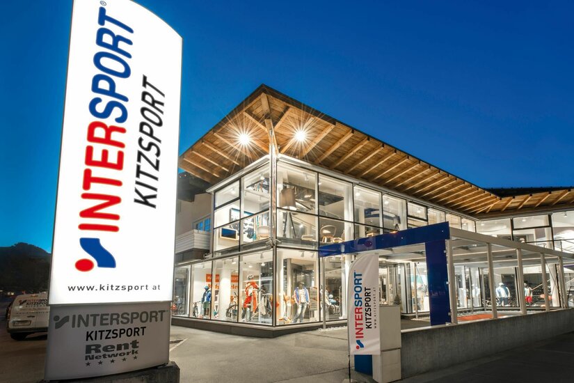 Intersport Kitzsport Filiale Beleuchtetes Sportgeschäft mit Glasfassade und großem Intersport-Werbeschild bei Dämmerung.