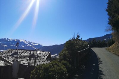 Asphaltierter Bergweg neben einem Hausdach mit Blick auf schneebedeckte Gipfel unter blauem Himmel.