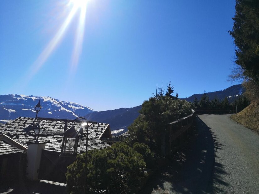Bergweg an der Astberg-Runde Asphaltierter Bergweg neben einem Hausdach mit Blick auf schneebedeckte Gipfel unter blauem Himmel.