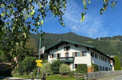 Ein charmantes Haus umgeben von Bäumen und grüner Natur. Im Hintergrund sind sanfte Berge und ein klarer blauer Himmel zu sehen.