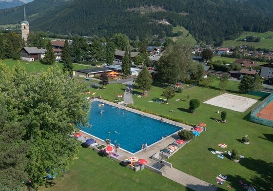 Freibad II Ein schöner Pool in einer ländlichen Umgebung, umgeben von grünen Wiesen und Bergen. Im Wasser sind Menschen beim Schwimmen, und es gibt Liegen mit Sonnenschirmen an den Rändern.
