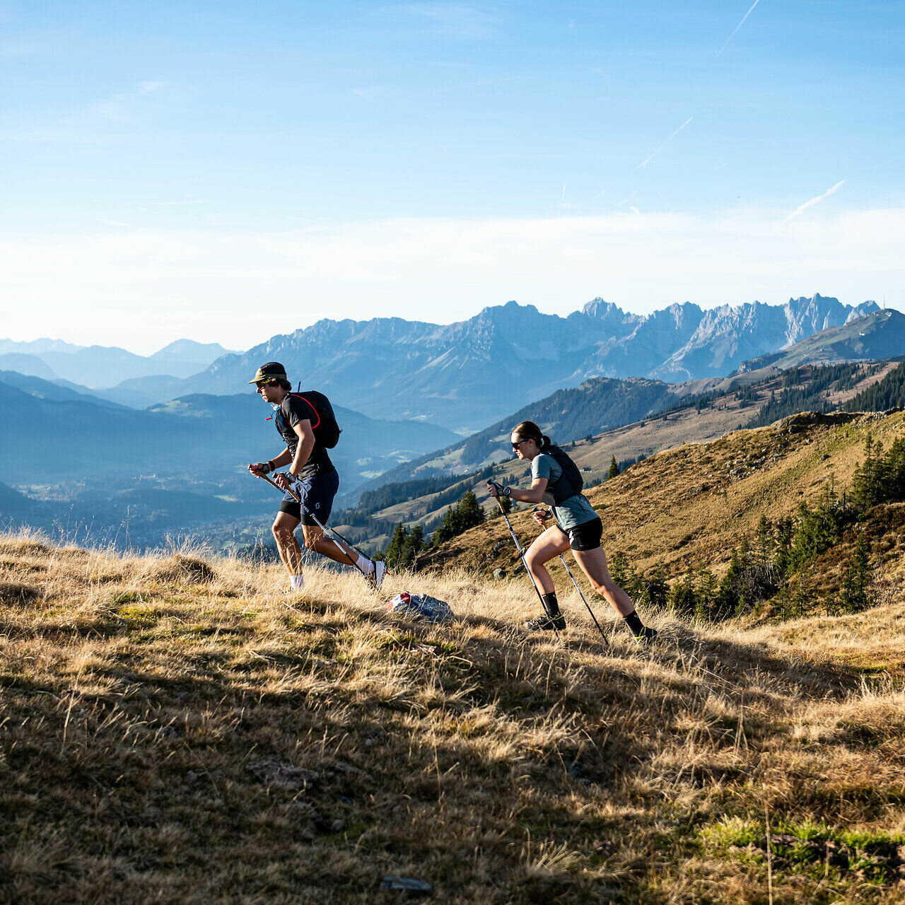 Zwei Trailrunner erklimmen einen hügeligen Pfad, umgeben von majestätischen Bergen und einer weiten, grünen Landschaft.