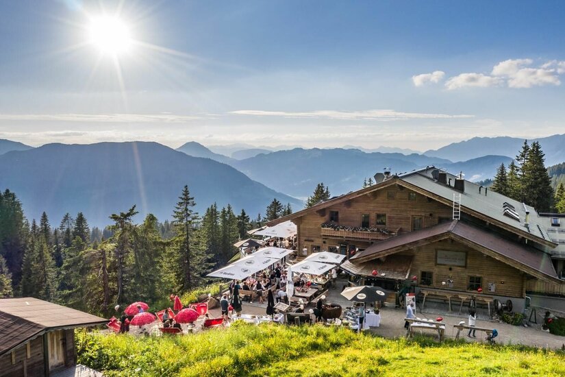 Berghütte aus Holz mit belebter Terrasse und Sonnenschirmen vor Gebirgspanorama bei Sonnenschein.