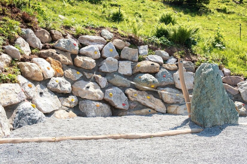 Künstliche Boulderwand aus Natursteinen mit bunten Klettergriffen auf einem Kiesplatz.
