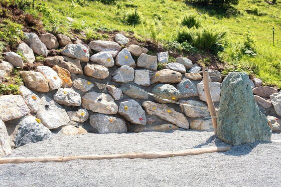 Künstliche Boulderwand aus Natursteinen mit bunten Klettergriffen auf einem Kiesplatz.