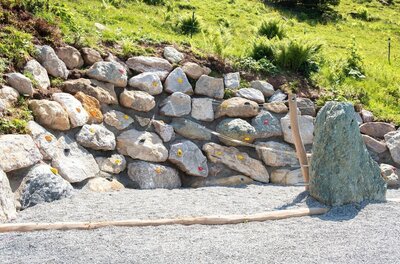 Künstliche Boulderwand aus Natursteinen mit bunten Klettergriffen auf einem Kiesplatz.