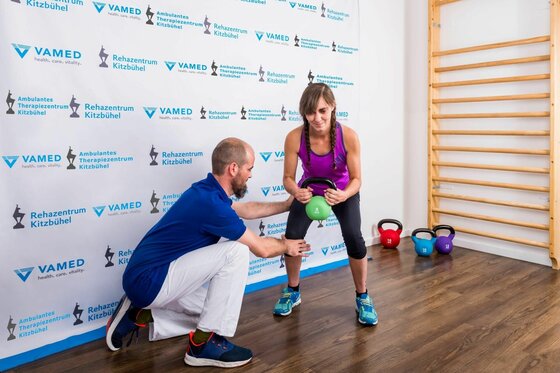 Therapie im Rehazentrum Kitzbühel Eine Frau trainiert mit einer Kettlebell unter Anleitung eines Therapeuten in einem hellen Raum.