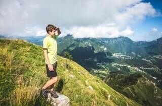 Ein Junge steht auf einem grasbewachsenen Berggipfel und blickt mit der Hand an der Stirn in die Ferne.