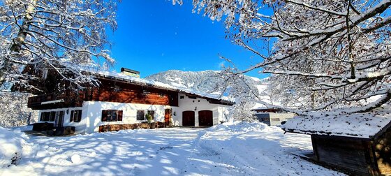 Außenansicht Villa Alpenblick im Winter Verschneites Holzhaus mit weißem Erdgeschoss vor bewaldeten Bergen unter blauem Himmel.