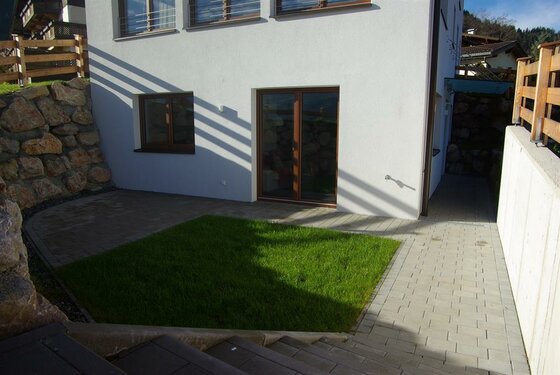 Gepflasterte Terrasse mit kleiner Rasenfläche vor einer weißen Hausfassade mit Glastür.