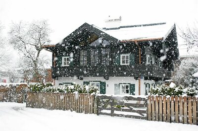 Verschneites zweistöckiges Holzhaus mit weißer Fassade und dunklem Balkon bei Schneefall.