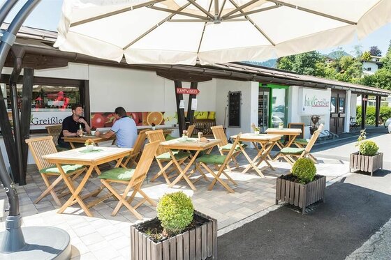 Außenbereich eines Cafés mit Holztischen, Stühlen und Sonnenschirm vor einem Gebäude.