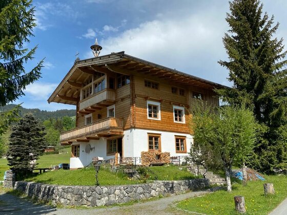 Dreistöckiges alpines Holzhaus mit weißem Erdgeschoss, Balkonen und Steinfundament vor Nadelbäumen.
