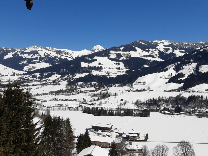 Verschneite Gebirgslandschaft mit bewaldeten Hängen und kleinen Siedlungen unter blauem Himmel.