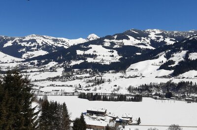 Verschneite Gebirgslandschaft mit bewaldeten Hängen und kleinen Siedlungen unter blauem Himmel.