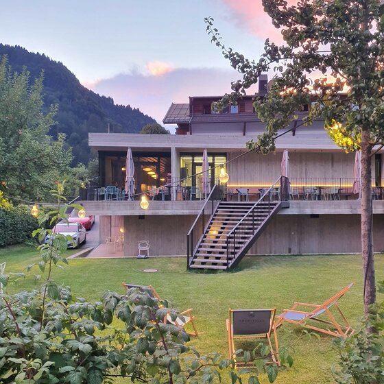 Modernes Gebäude mit Außenterrasse, Treppe und Liegestühlen auf einer Wiese vor bewaldeten Bergen.