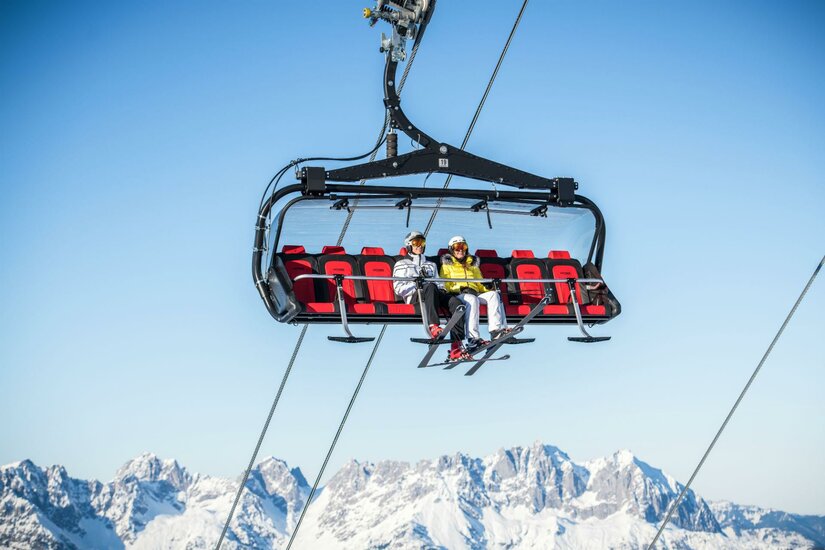 Skigebiet Ein Sessellift im Schnee mit zwei Personen, die hinauf in die Berge fahren. Im Hintergrund sind schneebedeckte Gipfel und ein klarer blauer Himmel zu sehen.