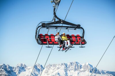 Skigebiet Ein Sessellift im Schnee mit zwei Personen, die hinauf in die Berge fahren. Im Hintergrund sind schneebedeckte Gipfel und ein klarer blauer Himmel zu sehen.