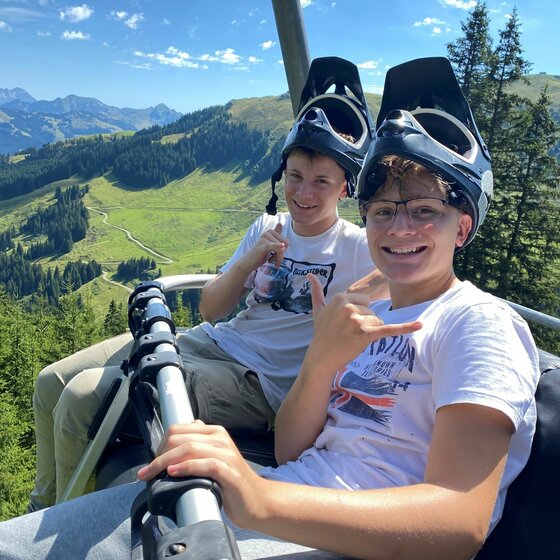 Mountainbiker im Sessellift Zwei Jugendliche mit Mountainbike-Helmen sitzen in einem Sessellift vor einer Bergkulisse.