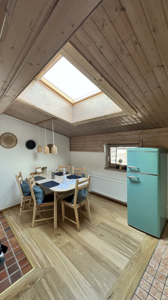 Eine gemütliche Küche mit einem großen Tisch und Holzstühlen. Ein blauer Kühlschrank steht neben einem Fenster mit Dachluke.