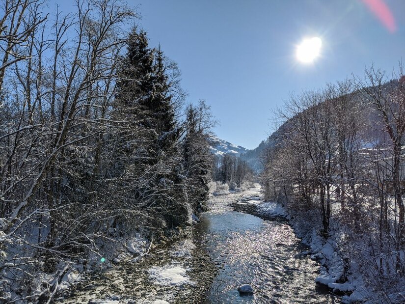 Winterlicher Flusslauf bei Malerndörfl Ein flacher Flusslauf zwischen verschneiten Bäumen unter blauem Himmel mit Sonnenschein.