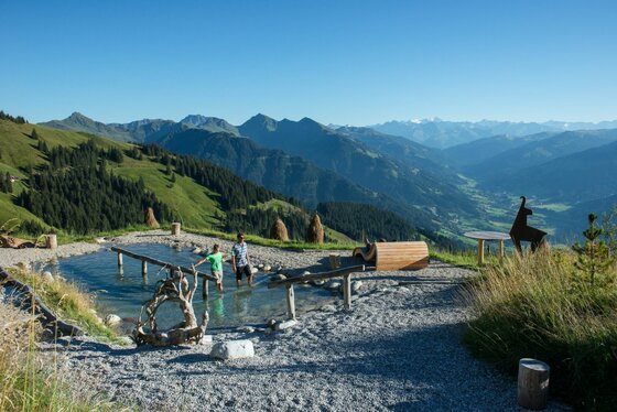 Kneippanlage an der Bichlalm Zwei Personen in einem Kneippbecken auf einem Bergplateau vor einem weiten Gebirgspanorama.