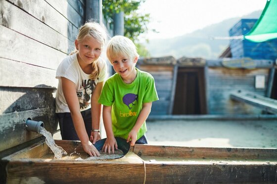 Kinder im Edelsteinpark Zwei Kinder spielen an einer hölzernen Wasser- und Sandrinne im Freien.